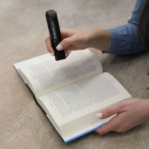 Hand using the OrCam Read device to read a book sitting on the ground.